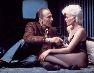 Scene from Brian De Palma’s Body Double (1984) with a woman in black lingerie talking to a man in a leather jacket on a bed in a Hollywood apartment high above Los Angeles, California.