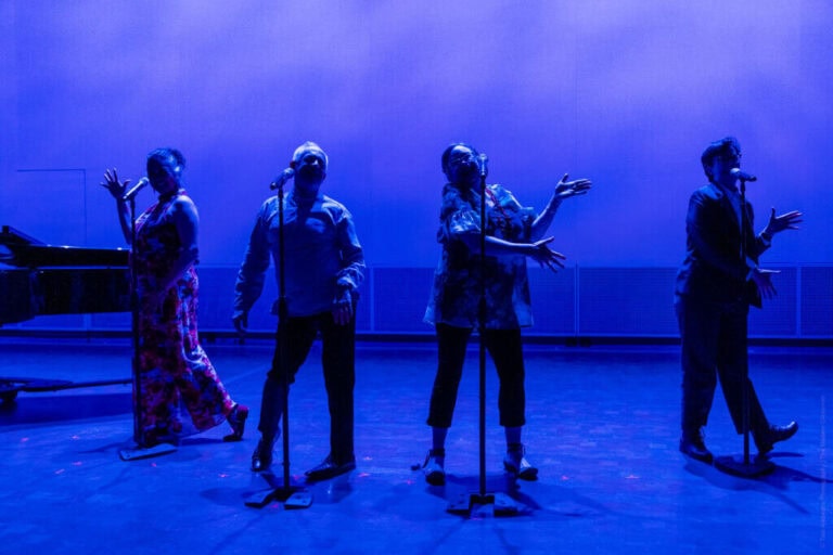 Four performers stand at microphones on a stage bathed in dramatic blue lighting during a Theatre Bay Area production