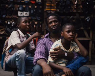 Sope Dirisu as Fola sits with his two young sons in front of a shoe vendor stall in Lagos in My Father's Shadow