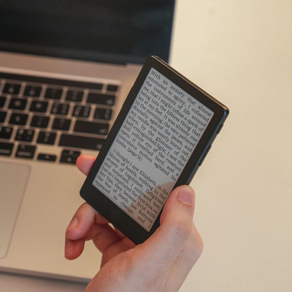 The XTEINK X4 Pro held in one hand in front of a laptop, showing a page of Mary Shelley Frankenstein on its 4.3-inch e-ink screen