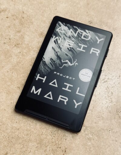 XTEINK X4 pocket e-reader resting on a concrete surface, its e-ink sleep screen displaying the cover art of Andy Weir's Project Hail Mary