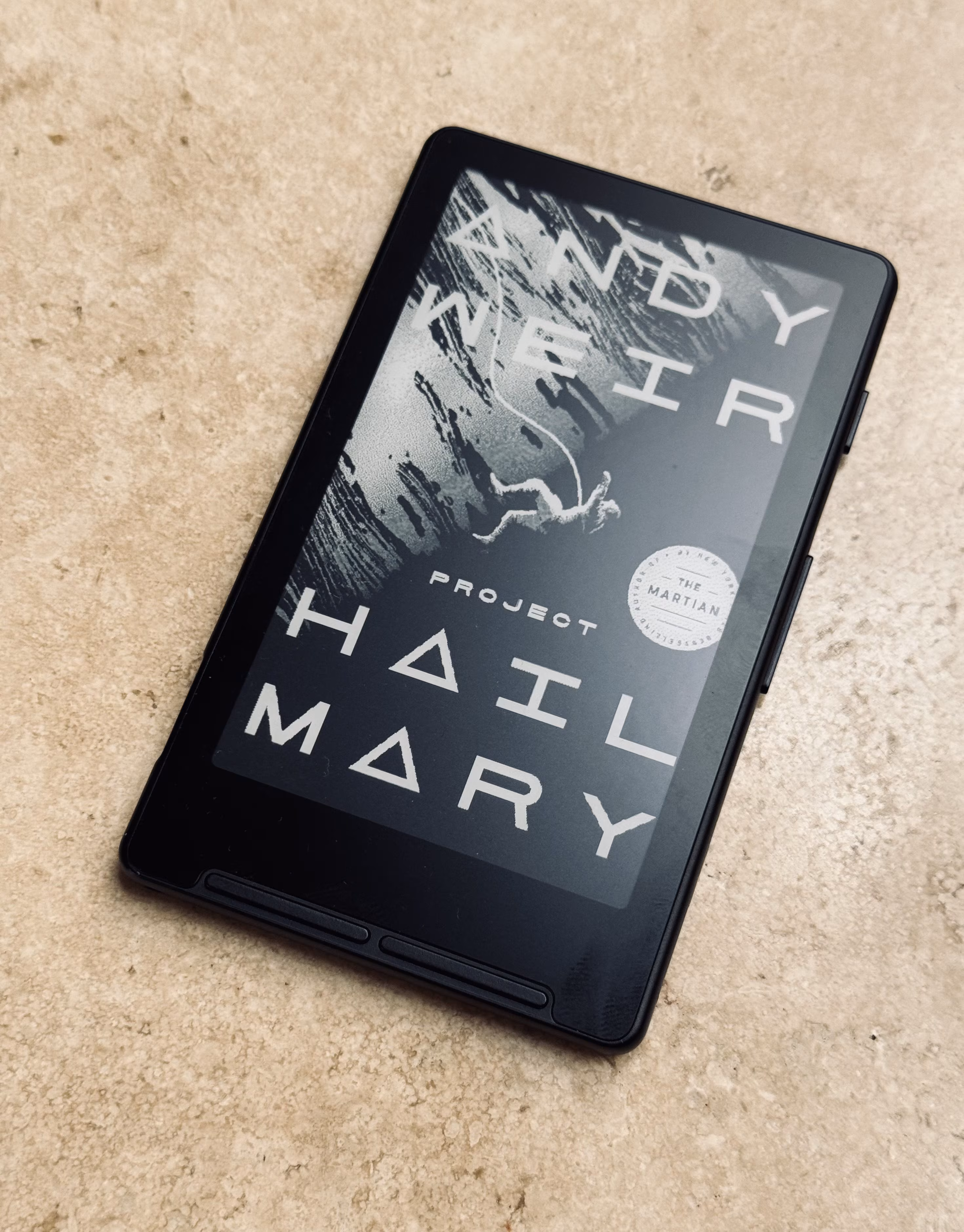 XTEINK X4 pocket e-reader resting on a concrete surface, its e-ink sleep screen displaying the cover art of Andy Weir's Project Hail Mary