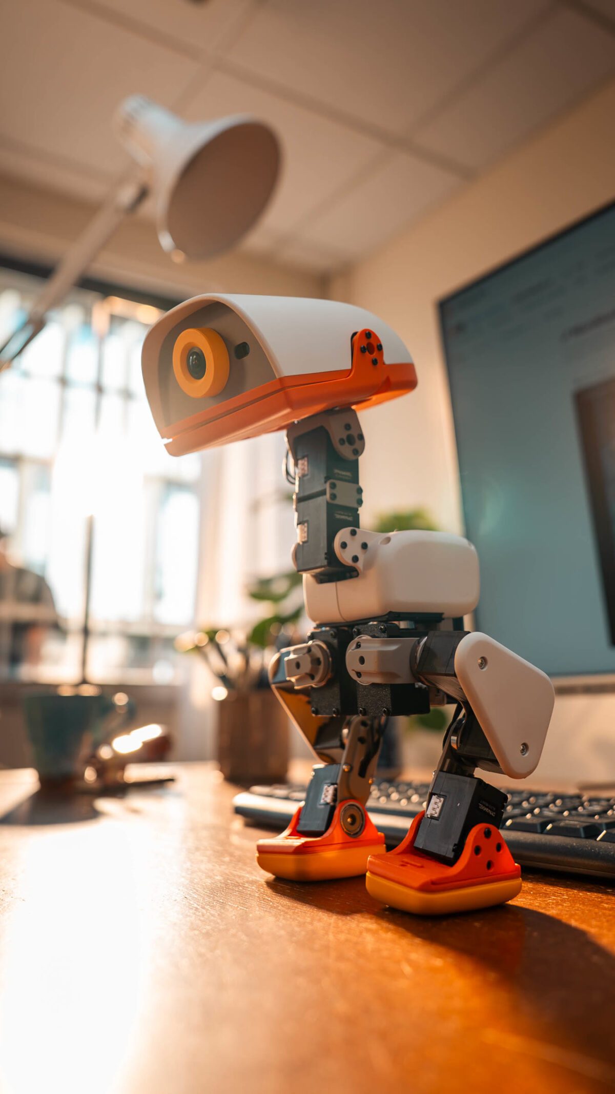 Close-up of a white and orange Microduck robot standing on a wooden desk in front of a keyboard and monitor