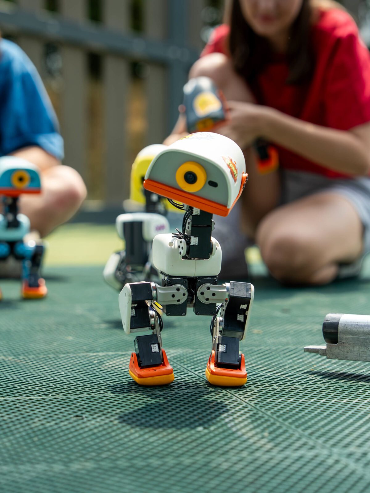 A Microduck robot walking toward the camera on an outdoor mat, with other Microducks and their owners in the background